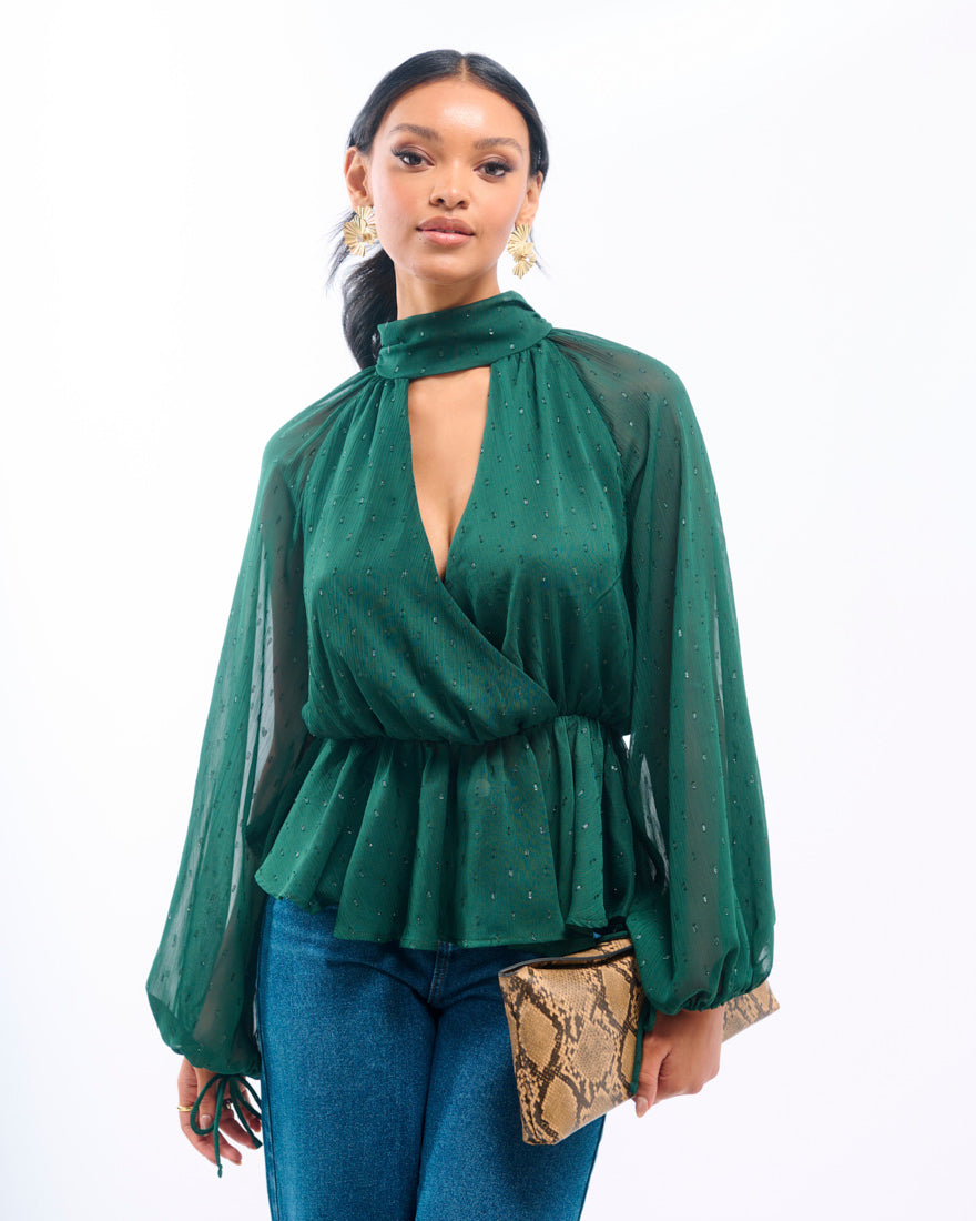 Woman wearing a green blouse with sheer sleeves and cut-out design, holding a snake skin clutch.
